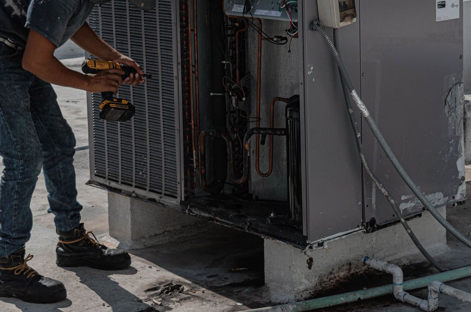 A worker in a bucket hat repairs an outdoor air conditioning unit on a rooftop.